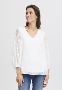 Weißes V-Ausschnitt-Bluse mit strukturiertem Blattmuster, langen transparenten Ärmeln und elastischen Bündchen; kombiniert mit blauen Jeans.