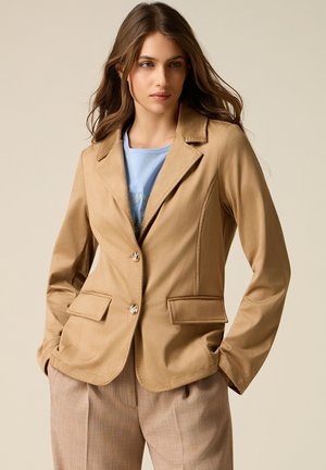 Mujer con blazer beige sobre una camisa azul claro con diseño de pedrería, combinada con pantalones marrón claro a cuadros, con las manos en los bolsillos.