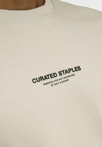 Beige sweatshirt med ribbet halsudskæring, der har sort trykt tekst "CURATED STAPLES" og "ESSENTIAL FOR ANY WARDROBE" på forsiden.