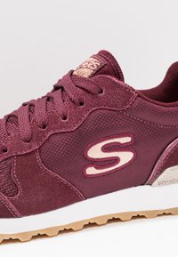 Burgundy athletic shoe with suede and mesh materials, featuring a textured surface and contrasting beige logo and accents. White midsole.