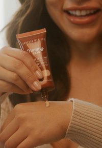 Bronsrör med "Yummy Skin" hudtoningskräm med en munstycke som dispenserar en ljusbeige produkt på ett handled. Slät textur, mjukt utseende och matt finish.