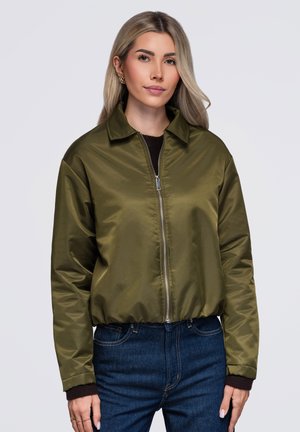 Femme aux longs cheveux blonds porte une veste bomber vert olive zippée sur un haut noir, associée à un jean bleu foncé sur un fond uni.