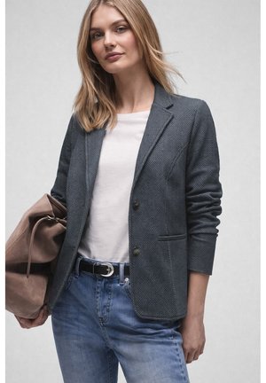 Kobieta ubrana w teksturowaną, ciemnoszarą marynarkę, białą koszulkę, niebieskie dżinsy, trzyma dużą brązową zamszową torbę na pasku.