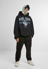 Felpa nera con logo blu, pantaloni cargo neri e sneakers crema. Gli accessori includono una sciarpa a motivi e occhiali da sole bianchi.