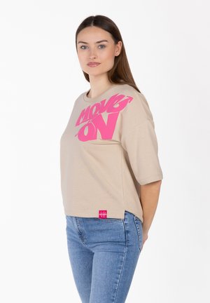 Kobieta o długich brązowych włosach nosi beżowy, oversize'owy T-shirt z różowym napisem "MOVE ON" oraz niebieskie dżinsy, stoi bokiem na białym tle.