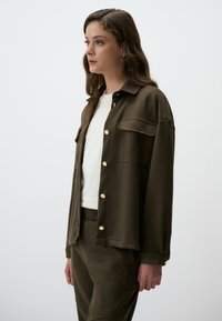 Giacca khaki a bottoni in morbido tessuto con tasca e bottoni dorati, abbinata a una maglietta bianca e pantaloni verde oliva coordinati.