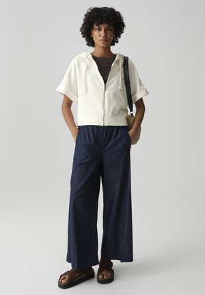 Donna con capelli ricci che indossa una felpa crema a maniche corte con cappuccio, pantaloni larghi scuri, sandali marroni e porta una borsa a tracolla beige.