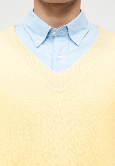 Ανοιχτό μπλε πουκάμισο με κουμπιά κάτω από ανοιχτό κίτρινο πλεκτό πουλόβερ με V λαιμόκοψη σε ένα άτομο με ουδέτερο φόντο.