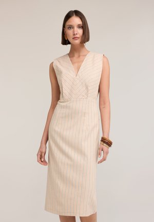 Femme aux cheveux courts bruns portant une robe beige sans manches avec de fines rayures verticales bleues et un décolleté en V, accessoirisée de bracelets en bois.