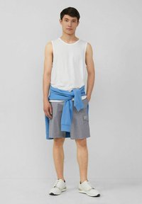 Weißes ärmelloses Hemd, blauer Pullover um die Taille gebunden, graue Shorts mit Taschen und weiße Sneakers. Einfaches Design und lässiger Stil.