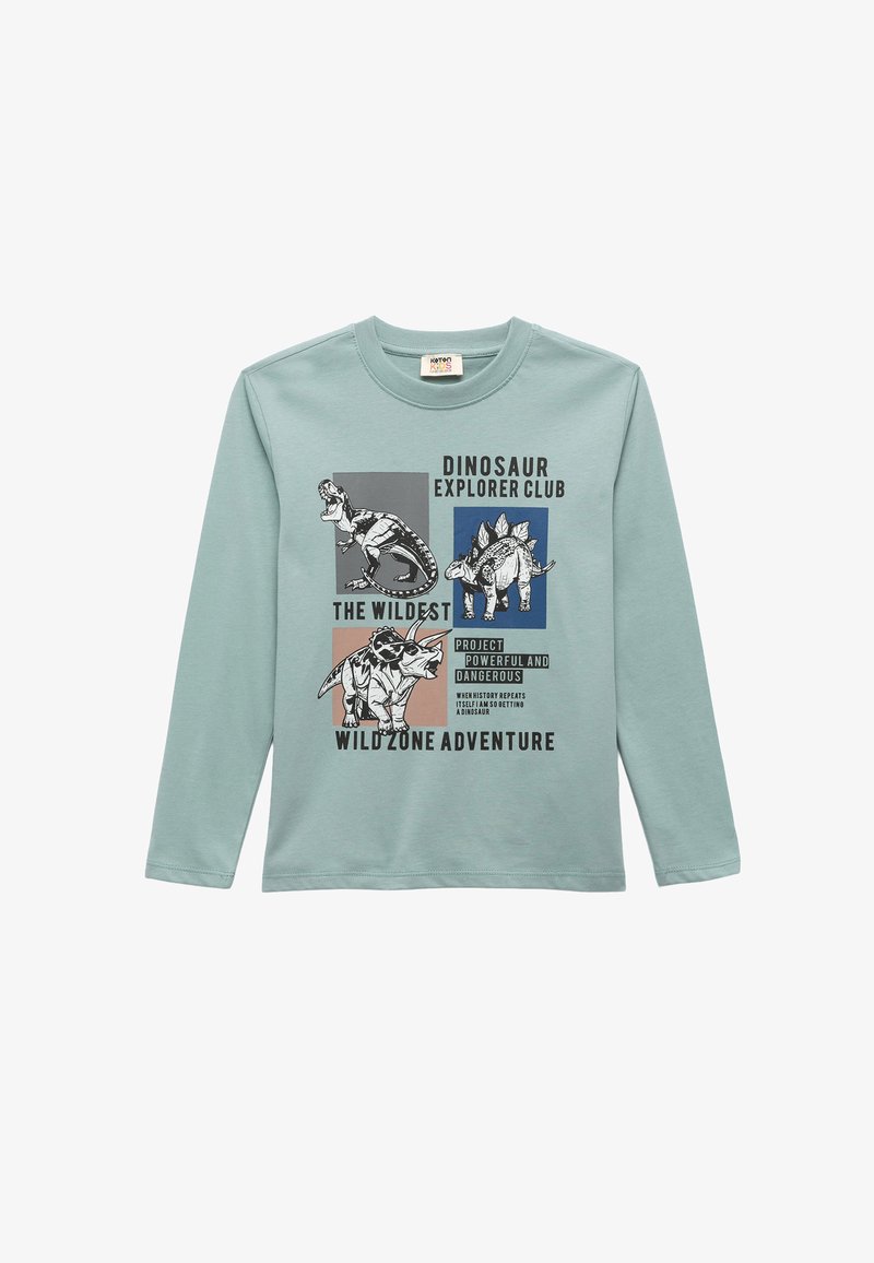 Camicia a maniche lunghe azzurro chiaro con grafiche di dinosauri e testo. I materiali sembrano morbidi; il design include quadrati colorati e caratteri in grassetto.