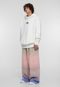 Felpa bianca oversize con tasca frontale e logo nero. Pantaloni a gamba larga con una sfumatura di colori dal rosa al blu. Semplici sneakers bianche.