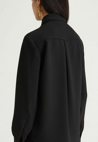 Blusa nera con colletto alto e maniche lunghe, realizzata in tessuto liscio, con pieghe sul retro e spacco agli orli delle maniche.