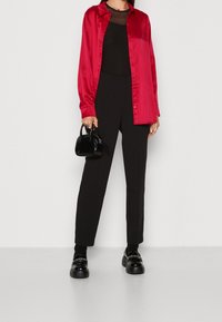 Rode satijnen overhemd met knopen over een zwarte mesh top, gecombineerd met zwarte op maat gemaakte broek en platform loafers. Kleine zwarte handtas met handvatten.
