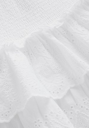 Tela blanca con textura fruncida en la parte superior, superpuesta con encaje con volantes que presenta patrones florales y pequeños recortes a lo largo del dobladillo.