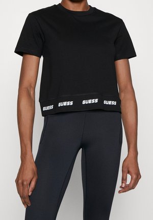 Personne portant un T-shirt noir court avec des logos GUESS le long de l'ourlet et des leggings sombres ajustés, debout devant un fond uni.