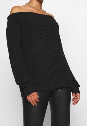 Vrouw die een zwarte, off-shoulder grofgebreide trui en zwarte leren broek draagt, gecompleteerd met een gouden ketting.