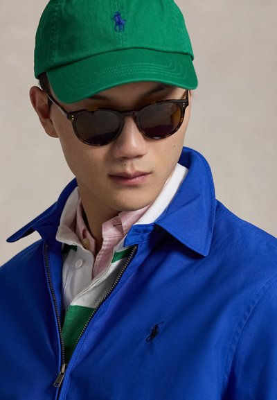 Μπλε μπουφάν με φερμουάρ και μαύρο λογότυπο, φορεμένο πάνω από λευκή πόλο με πράσινες ρίγες και ροζ γιακά. Πράσινο καπέλο και καφέ γυαλιά ηλίου.