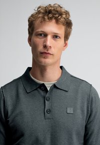 Dunkelgrau Polohemd mit Kragen, einer Knopfleiste mit drei Knöpfen und einem kleinen Logopatch. Glatte Textur, taillierte Passform.