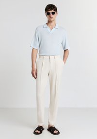 Mann trägt ein hellblaues Strick-Polo, cremefarbene, gefaltete Hose, schwarze geflochtene Sandalen und Sonnenbrille, steht vor einem schlichten weißen Hintergrund.