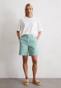 Leichtes weißes Baumwoll-T-Shirt mit einer Tasche, kombiniert mit mintgrünen Baumwollshorts. Getragen mit beigen, gewebten Plateausandalen.
