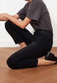 Vaqueros negros de talle alto y corte ajustado, con bolsillos delanteros y cremallera, combinados con una camiseta gris y zapatos de tacón negros sobre un suelo de madera.