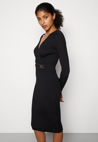 Zwart gebreid jurk met lange mouwen, diepe V-hals, geribbelde textuur, getailleerde silhouet en een contrasterende riem met een ronde gesp.