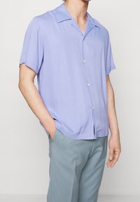 Camicia a maniche corte di colore viola chiaro con colletto, caratterizzata da una texture liscia e una vestibilità ampia. I bottoni sono visibili davanti.