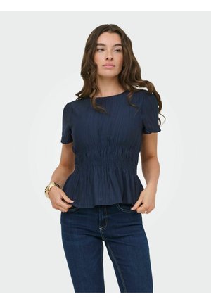 Femme aux longs cheveux ondulés portant un haut smocké bleu marine à manches courtes et un jean bleu foncé, debout devant un fond uni.