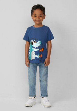 Blaues Baumwoll-T-Shirt mit einem strukturierten grünen Dinosaurier-Design mit Sonnenbrille und Flammen, kombiniert mit hellen Jeans und weißen Turnschuhen.