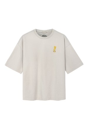 Jasnobeżowy, oversize T-shirt z krótkimi rękawami, z małą żółtą grafiką i tekstem na lewej stronie klatki piersiowej.