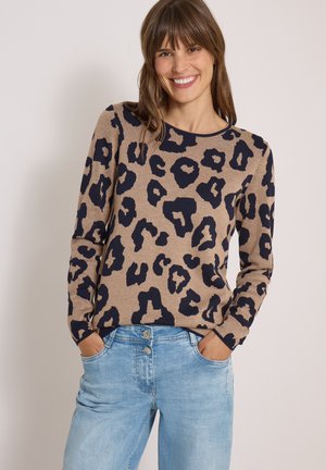 Femme souriante portant un pull beige avec des taches léopard noires et un jean bleu clair, les mains dans les poches sur un fond uni.