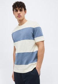 Νεαρός άνδρας με σγουρά μαλλιά, φορώντας κοντομάνικη μπλούζα με φαρδιές ρίγες σε μπλε και άσπρο και σκούρο παντελόνι, όρθιος με το ένα χέρι στην τσέπη.