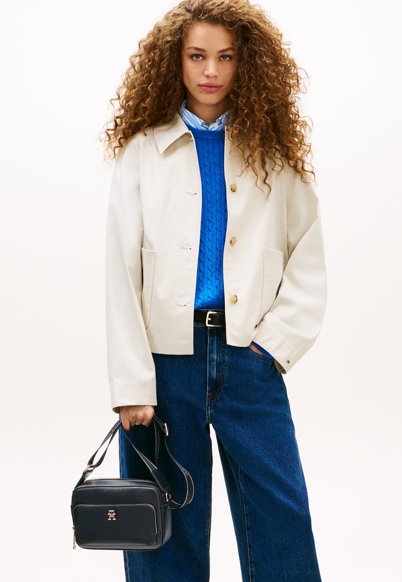 Tânără cu păr creț poartă o jachetă bej peste un pulover albastru și o cămașă în dungi, ținând o geantă neagră cu logo auriu.