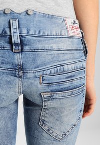 Bledé modré džíny s vysokým pasem, které mají dvě zadní kapsy a přišitý textilní štítek na pase. Texturovaná džínovina s mírným opotřebením.