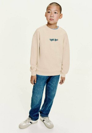 Sweatshirt beige avec le texte "PENSER GRAND !", col bleu clair, associé à un jean bleu et des baskets blanches avec des accents gris.