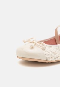 Ballet flat in un crema chiaro con punta in vernice lucida e tomaia in tessuto intrecciato, accentuata da un dettaglio a fiocco decorativo nella parte anteriore.