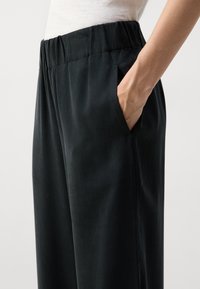 Schwarze, leichte, weit geschnittene Hosen mit elastischem Bund und Seitentaschen. Glatte Textur mit dezenter Satinierung und minimalistischen Design.