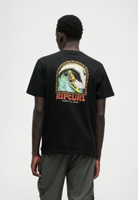 T-shirt noir avec un graphique coloré représentant un squelette surfant sur une vague, avec le texte "RIPCURL" et "SHRED TILL DEAD" en dessous du design.