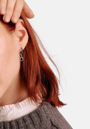 Rothaarige Person mit silbernem und schwarzem Edelstein-Tropfenohrring berührt das Haar, trägt einen grauen Strickpullover und ein weißes Hemd mit gerafftem Kragen.