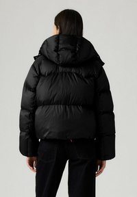 Vrouw in een zwarte gewatteerde jas met capuchon, gezien vanaf de achterkant, in combinatie met zwarte jeans tegen een effen lichte achtergrond.