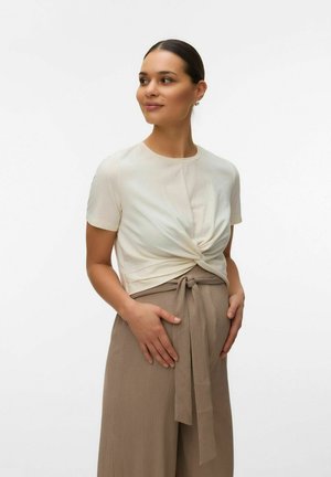 Femme enceinte avec un haut crème à manches courtes noué et un pantalon beige taille haute, les mains reposant sur le ventre, regardant vers la droite.