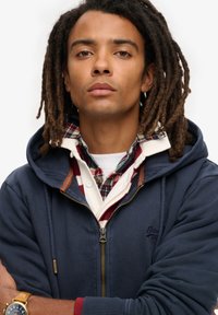 Marine blauwe zip-up hoodie met beige accenten, laag over een rood geruit overhemd. Opmerkelijke details zijn een logo en een zichtbare rits.