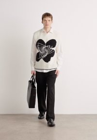 Pulover fără mâneci pentru bărbați, de culoare crem, tricotat, cu un mare design abstract negru. Asortat cu o cămașă albă, pantaloni negri și o geantă neagră.