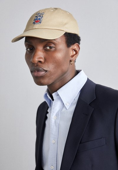 Casquette beige avec un motif graphique brodé multicolore, en coton doux, avec une visière incurvée. Portée avec une chemise bleu clair à boutons et une veste sombre.