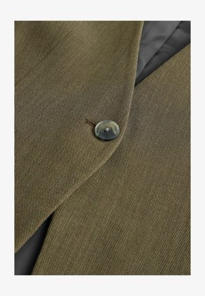 Brūnas tekstūras audums ar tuvplānu skatu uz pogu. Poga ir tumša un apaļa, piešūta pie pielāgota žaketes apkakles.