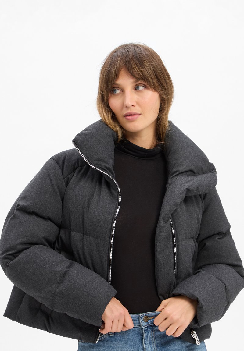Graue Puffjacke mit hohem Kragen, gestepptem Muster, Frontreißverschluss und weiten Ärmeln. Über einem schwarzen Oberteil getragen und mit blauen Jeans kombiniert.