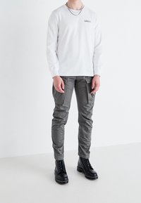Person in weißem Levi's Langarmhemd, grauen Cargo-Hosen, schwarzen Schnürstiefeln und einer silbernen Kettenkette, der vor einem einfarbigen weißen Hintergrund steht.