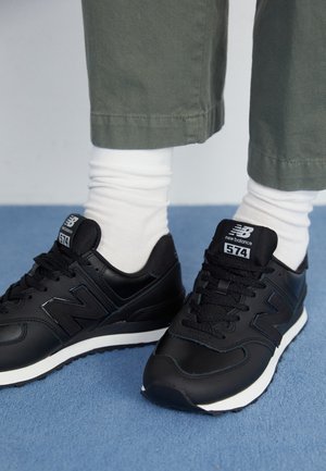 Stopy w czarnych butach sportowych New Balance 574 z białymi podeszwami, białymi skarpetkami i oliwkowozielonymi spodniami stojące na niebieskiej podłodze.