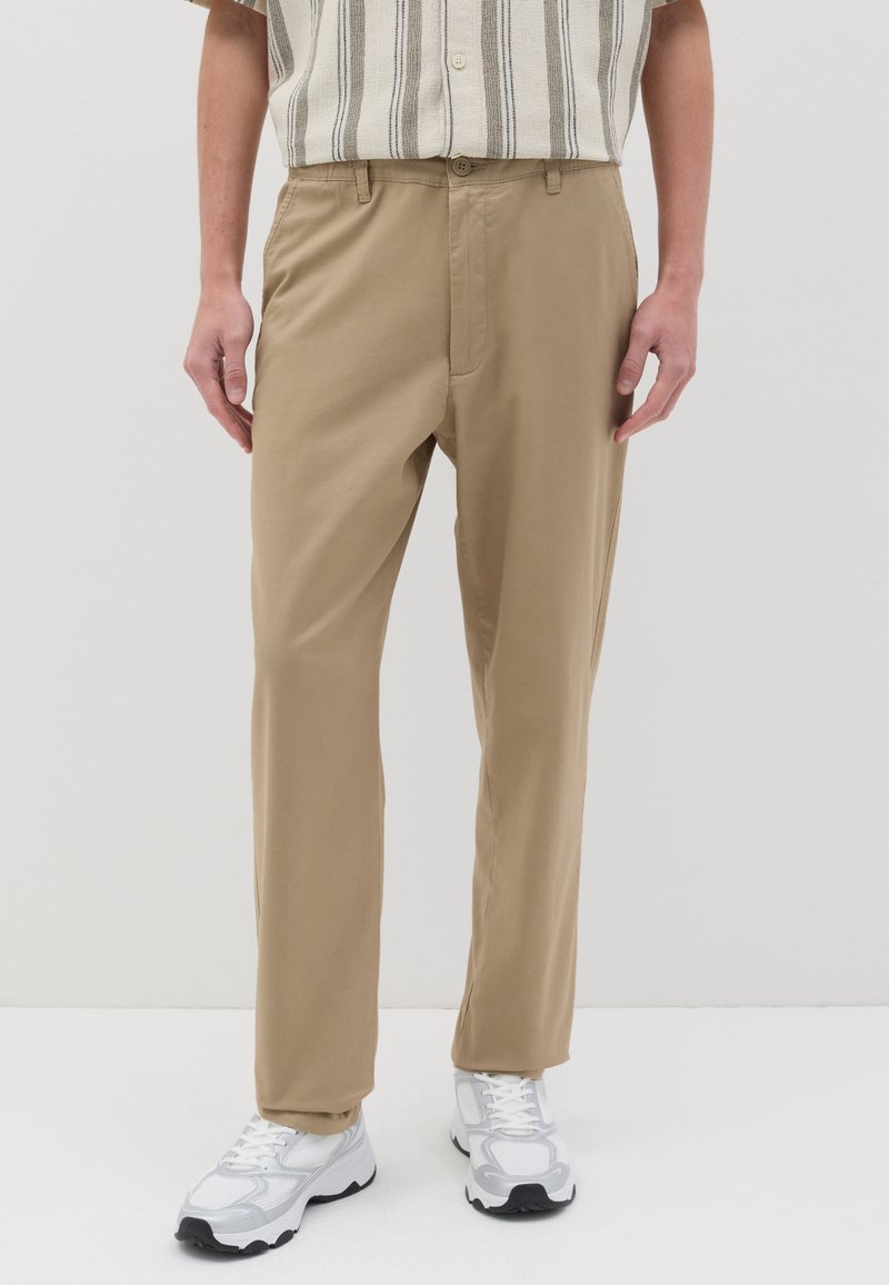 Pantalon kaki à coupe droite, avec fermeture à bouton et poches latérales, associé à des baskets blanches. Tissu lisse et couleur neutre.